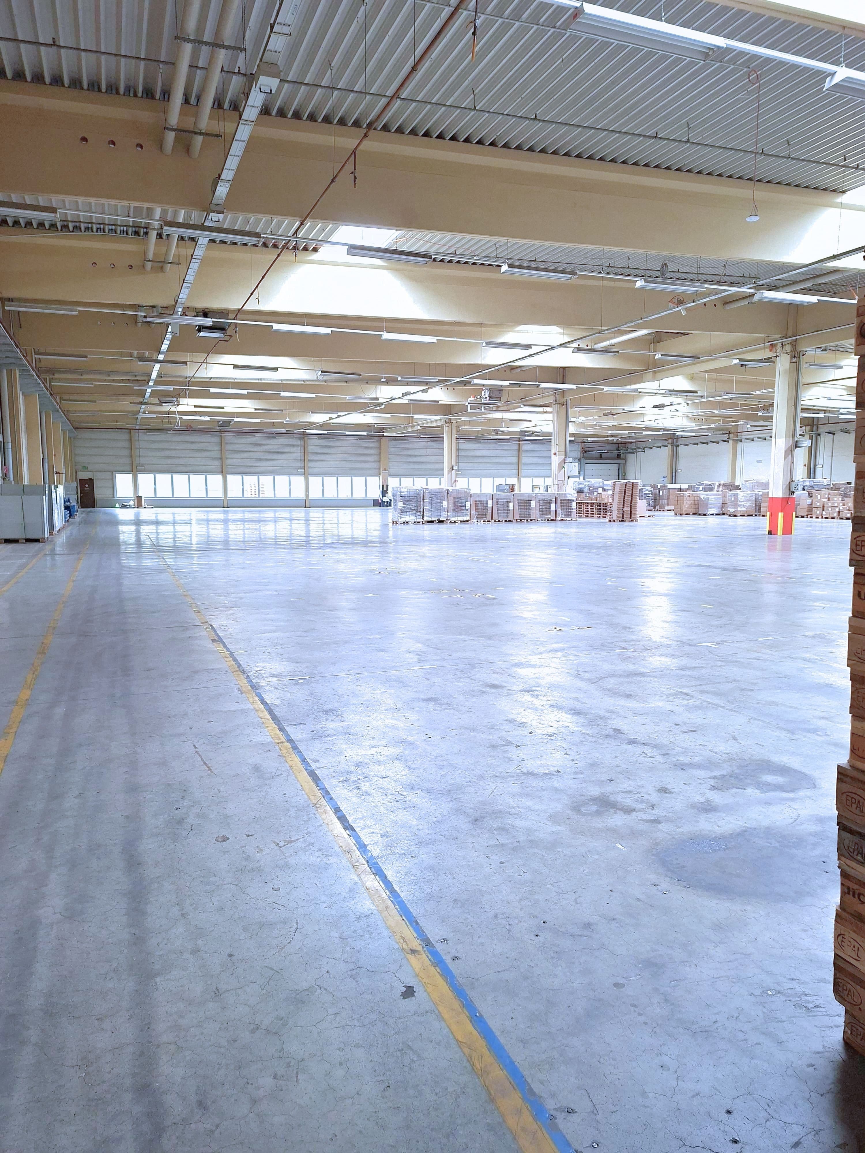 Weitläufige Lagerhalle mit glänzendem Betonboden, gelben Markierungslinien und im Hintergrund gestapelten Paletten mit Waren.