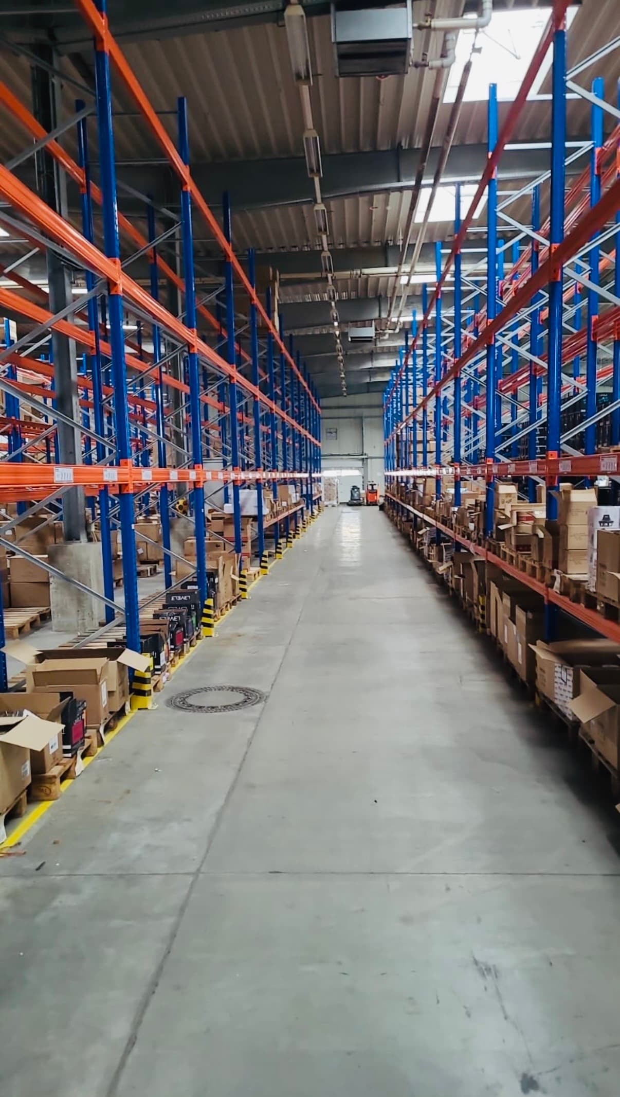 Langer Gang in einem Lagerhaus mit hohen blau-orangen Regalen und vielen gestapelten Kartons.