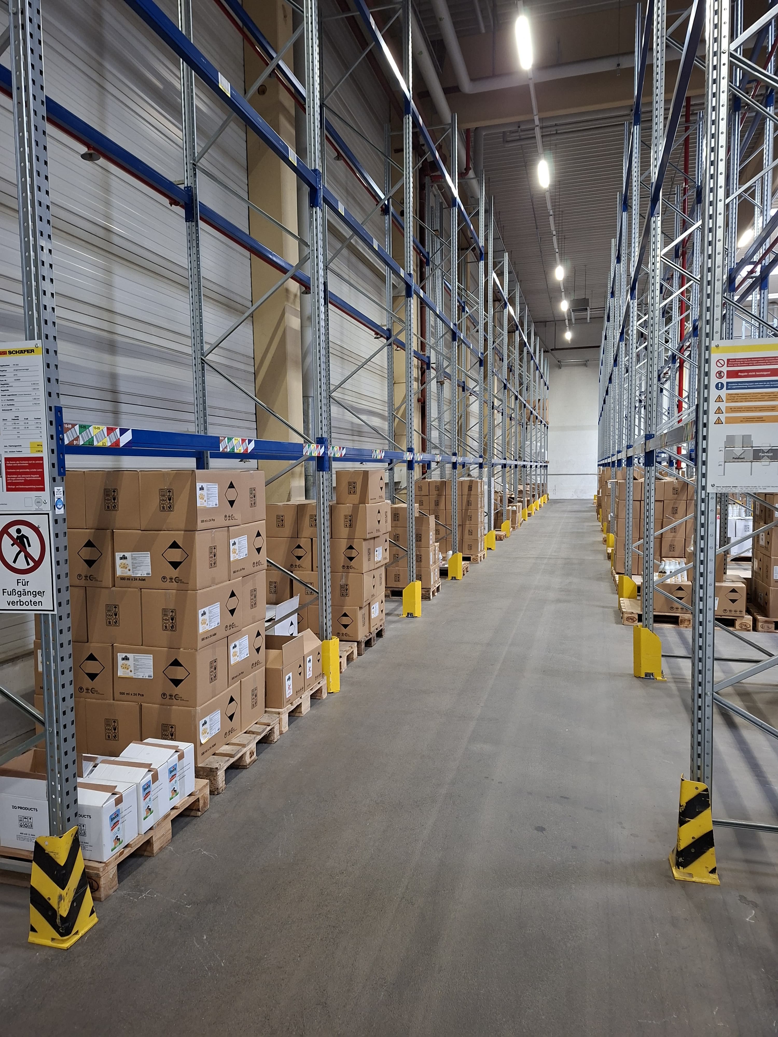 Ein langer Gang in einem Lagerhaus mit hohen Metallregalen und auf Paletten gestapelten Kartons.