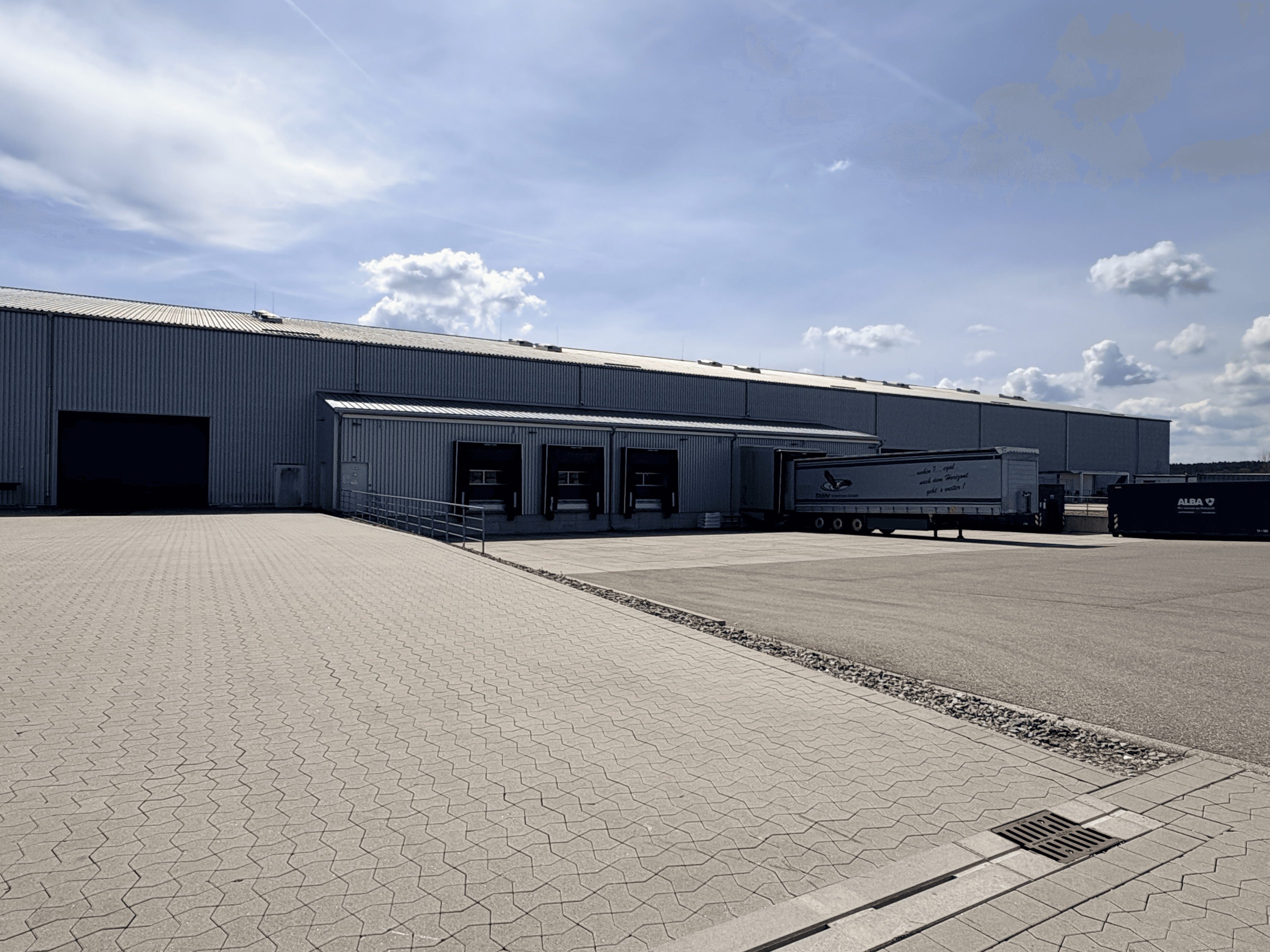 Große graue Industriehalle mit Laderampen und einem LKW-Anhänger auf einem gepflasterten Vorplatz.