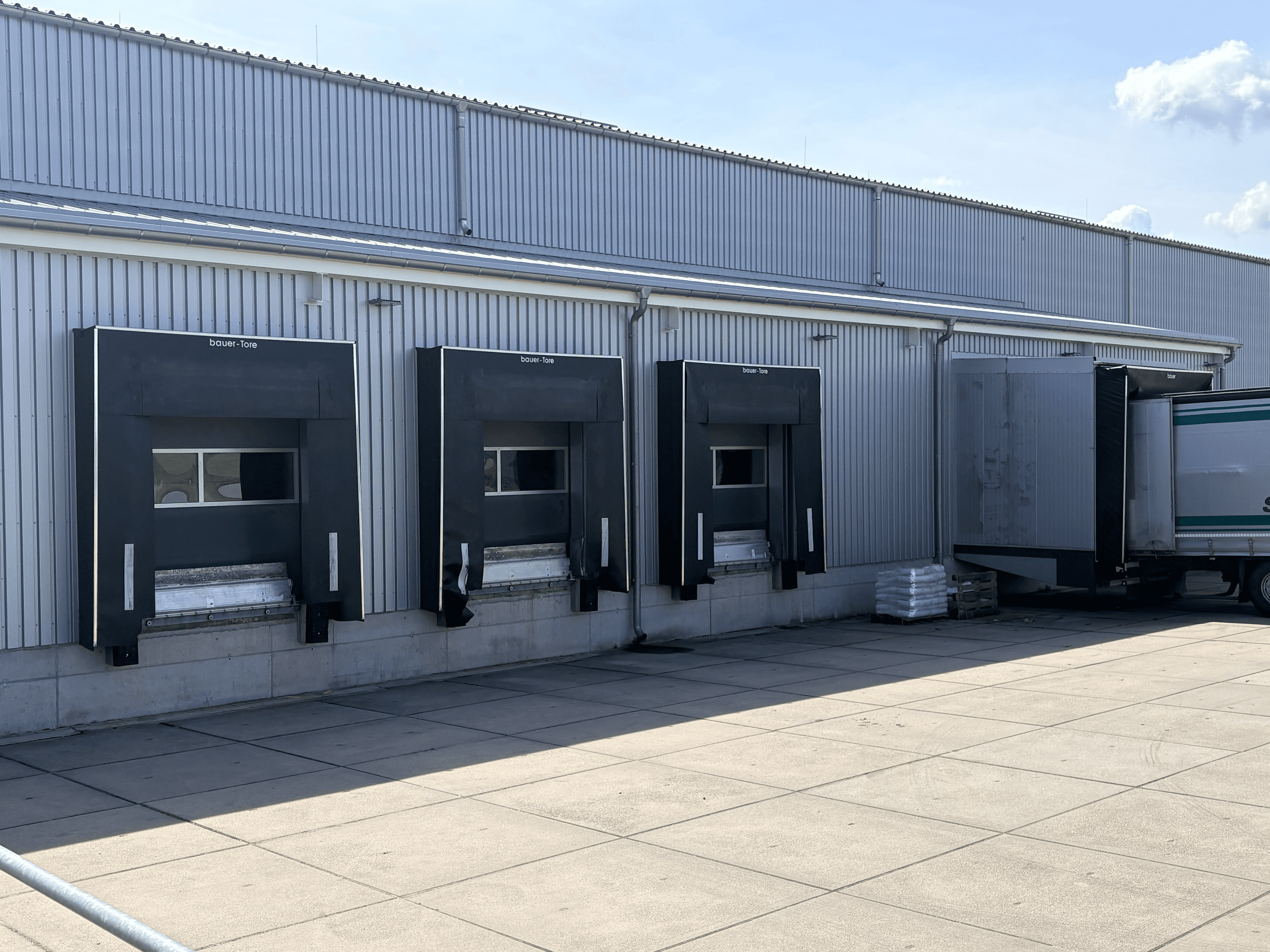 Graue Lagerhalle mit mehreren Laderampen und einem angedockten LKW vor einer modernen Wellblechfassade.